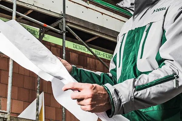  A man holding a blueprint of a steel structure is standing in front of a building that has scaffolding built around the building.