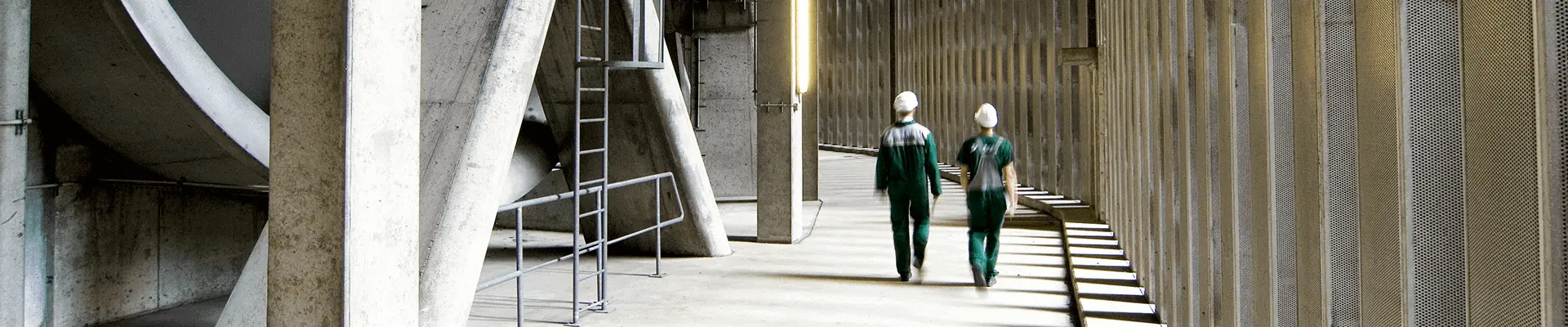 Two people walk through a corridor built entirely of concrete with a high ceiling and pillars.