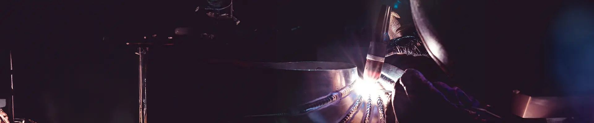 Närbild på en svetsare i mask som svetsar ihop olika delar av ett tjocks rör i titan.