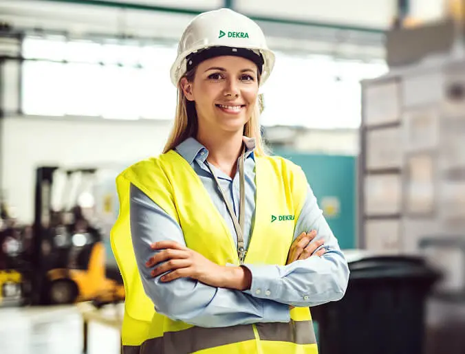 Kvinna med armarna i kors, iförd gul väst och vit hjälm med grön DEKRA-text, står och tittar mot kameran. Kvinna med armarna i kors, iförd gul väst och vit hjälm med grön DEKRA-text, står och tittar mot kameran.