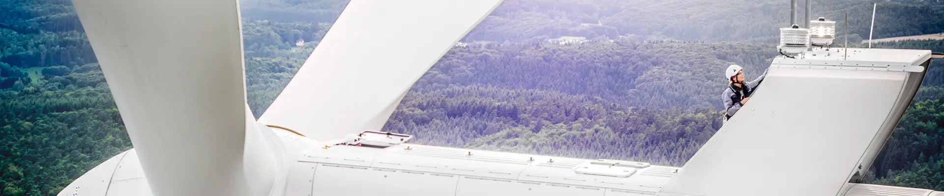 A technician in safety gear inspects a wind turbine high above a forested area, showcasing the maintenance work involved in renewable energy. A technician in safety gear inspects a wind turbine high above a forested area, showcasing the maintenance work involved in renewable energy.