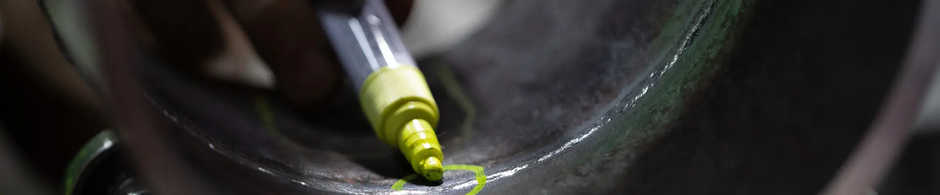 A person's white-gloved hand holds a yellow marker pen and draws a circle around a damaged area in a steel pipe.