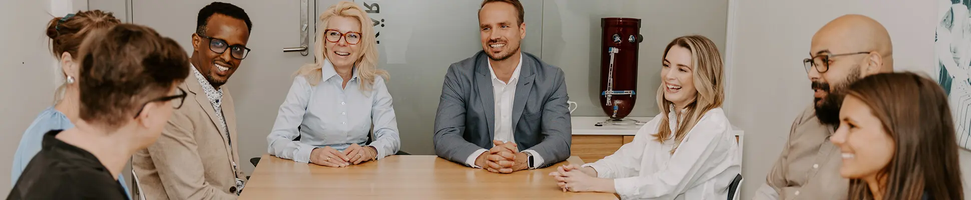 Flertalet medarbetare som sitter vid ett bord i ett konferensrum, ler och tittar på varandra.