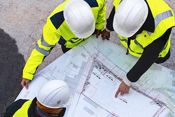 A diverse group of individuals wearing bright yellow vests stand together, engaged in a construction drawing.