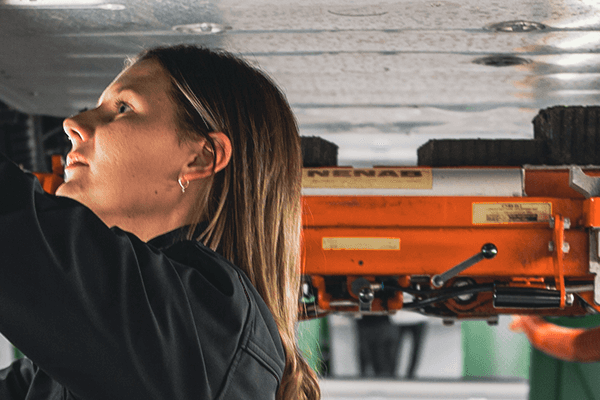 A woman carrying out a car inspection stands under a car that is raised, she inspects the car from below and shines a flashlight.