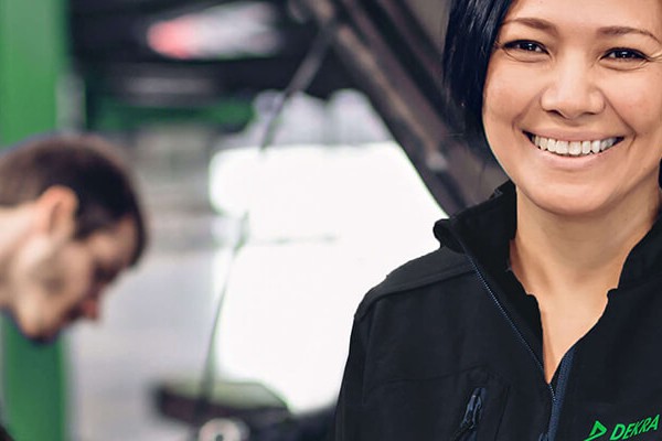 A woman in black work clothes stands in front of a background in the form of a car workshop with a man working on a picture engine with the hood open.