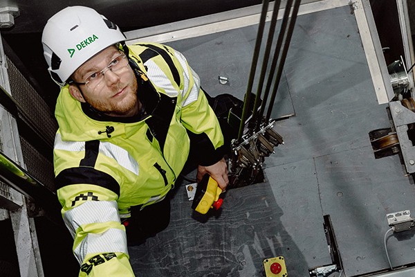 A man in a yellow jacket stands on the roof of an elevator inside an elevator shaft and looks up into the elevator shaft.