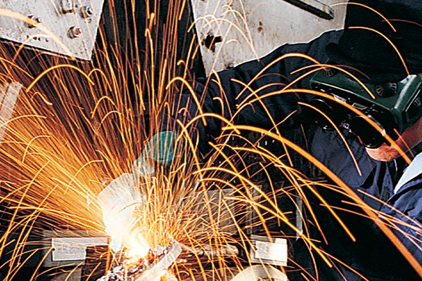 A man wearing protective gear is welding metal pieces together in a workshop, sparks flying from the welding torch.