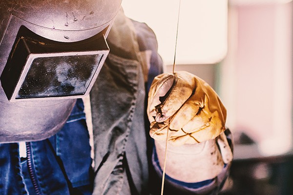 A man in protective equipment such as a helmet and jacket stands over an object to be welded in an industrial factory.