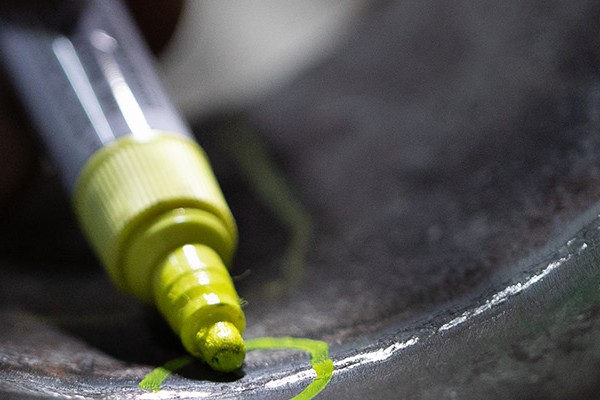 A person's white-gloved hand holds a yellow marker pen and draws a circle around a damaged area in a steel pipe.