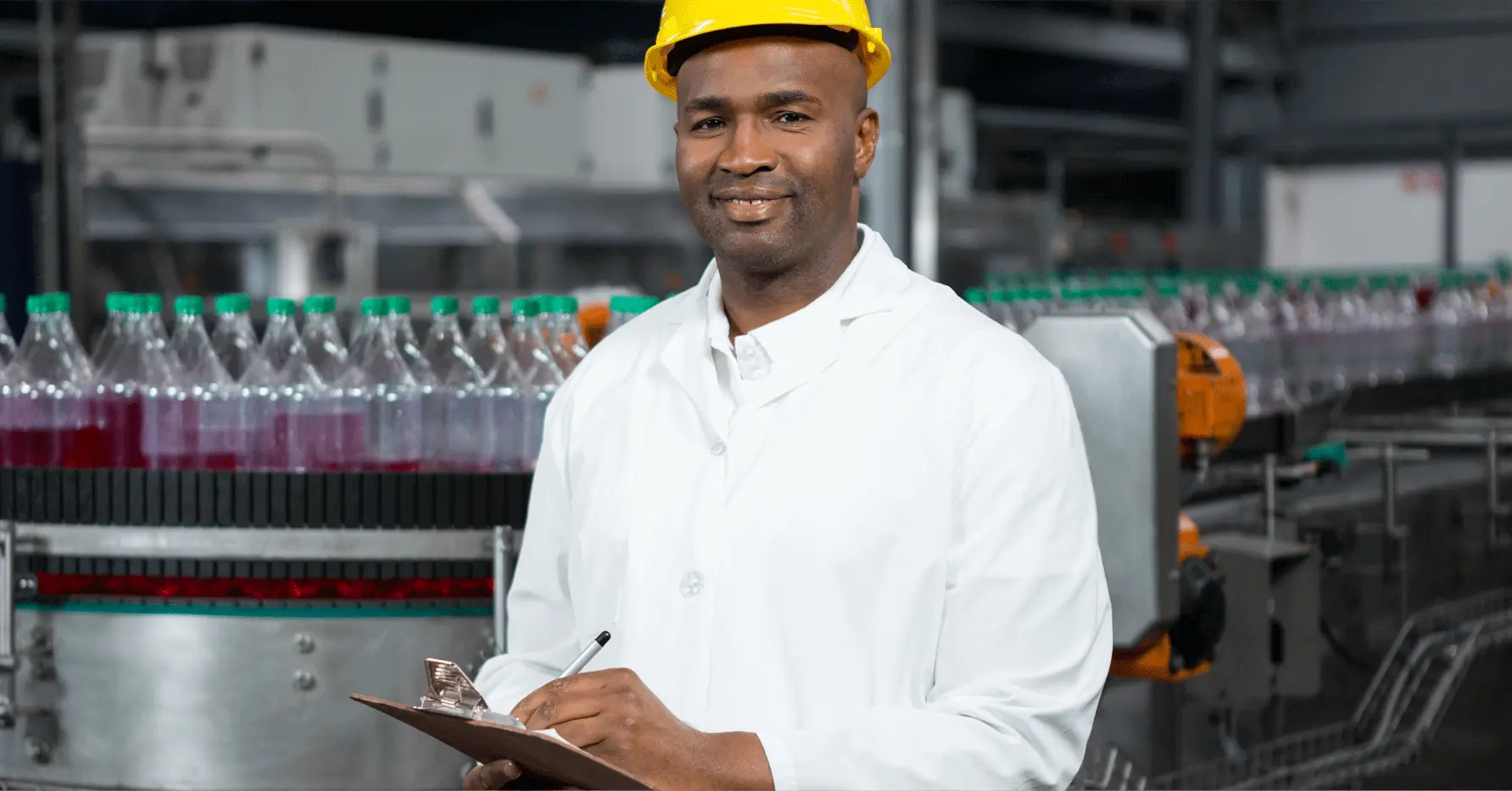 A factory worker wearing a yellow helmet and a white shirt. Behind him is a bottle assembly line. A factory worker wearing a yellow helmet and a white shirt. Behind him is a bottle assembly line.